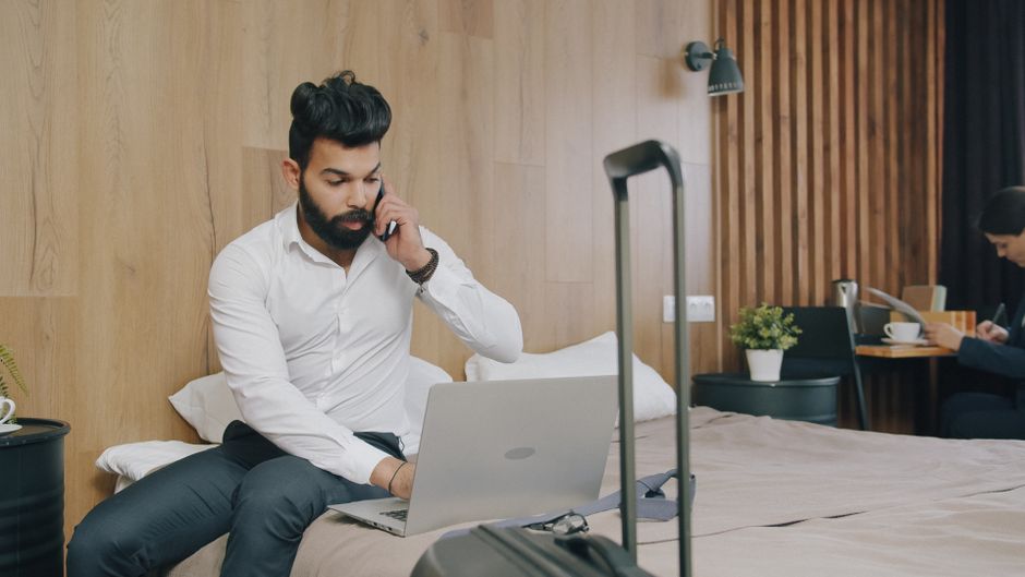 Businessman on laptop and phone in hotel room with luggage, working remotely.