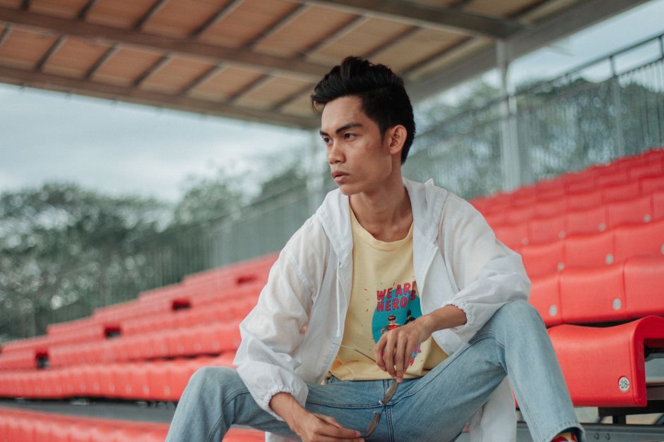 Young man posing casually on red stadium seats wearing denim jeans and a white jacket.