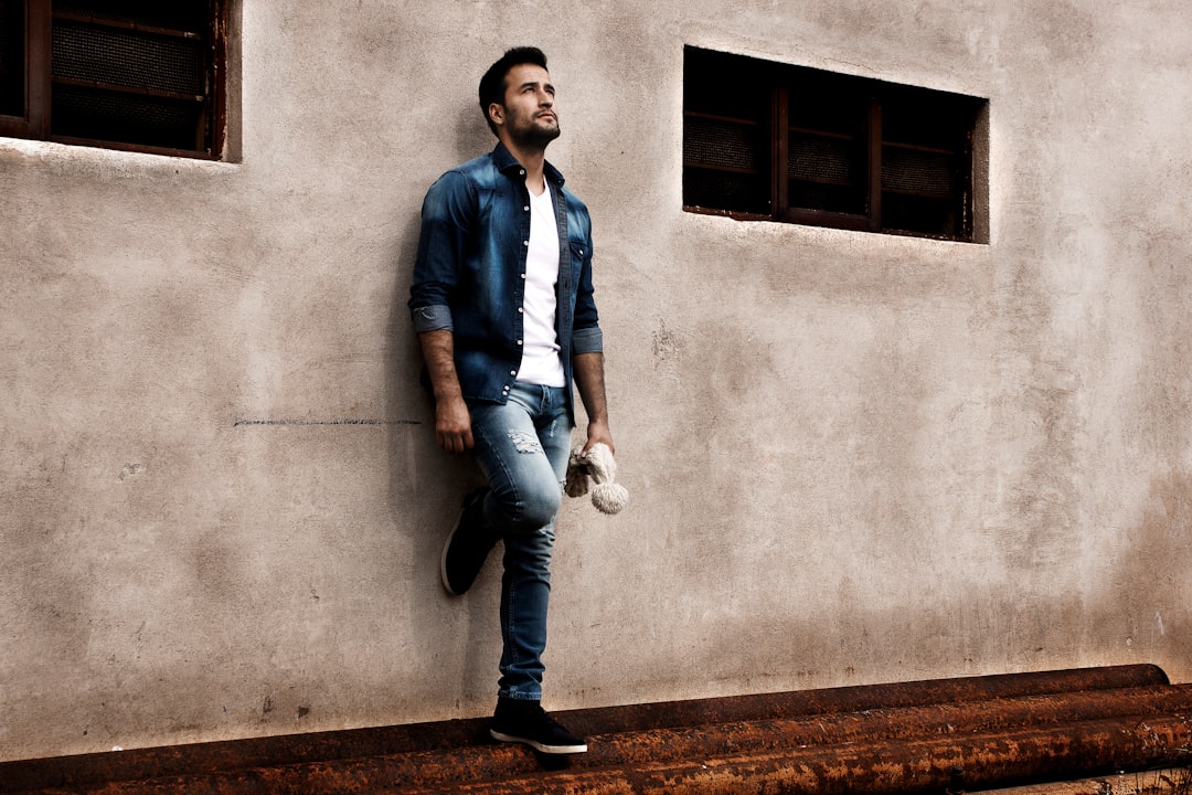 Man in blue dress shirt and blue denim jeans standing beside gray concrete wall during daytime